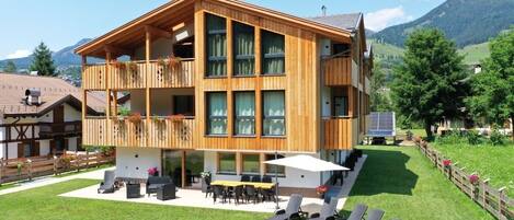 A two-story wooden building with a balcony, outdoor seating, and a view of mountains.