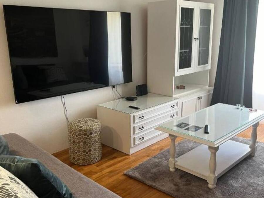 A modern living room with a flat-screen TV, a glass-top dining table, a white cabinet, and a patterned ottoman.