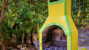 A green and yellow outdoor grill with a brick interior, surrounded by trees and a fence. - Nirvana Apartments & Auto Camp (Kumbor)