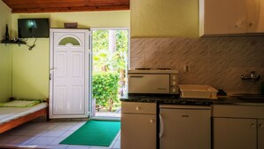A compact kitchen with a built-in oven, a refrigerator, and a microwave. There's a small dining area with a bench and a green mat. A white door leads to a balcony with a view of greenery. - Nirvana Apartments & Auto Camp (Kumbor)