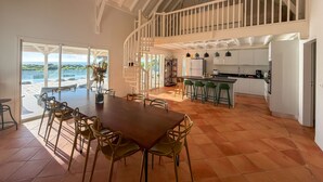 Ein geräumiger Essbereich mit großem Tisch und Stühlen, eine Küche mit weißen Schränken und Blick auf das Meer durch große Fenster.