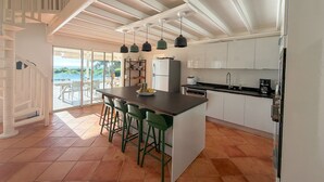 Eine moderne Küche mit einer zentralen Kochinsel, Barhocker, einem Kühlschrank und Blick ins Freie.