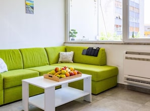 Un sofá verde, una mesa blanca con una cesta de fruta y un radiador.