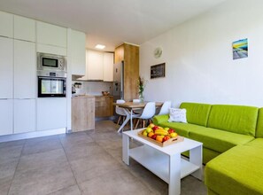 Una cocina moderna con armarios blancos, microondas y horno empotrado. Hay un comedor con una mesa y sillas de madera, y un sofá verde con una cesta de frutas sobre una mesa de centro blanca.