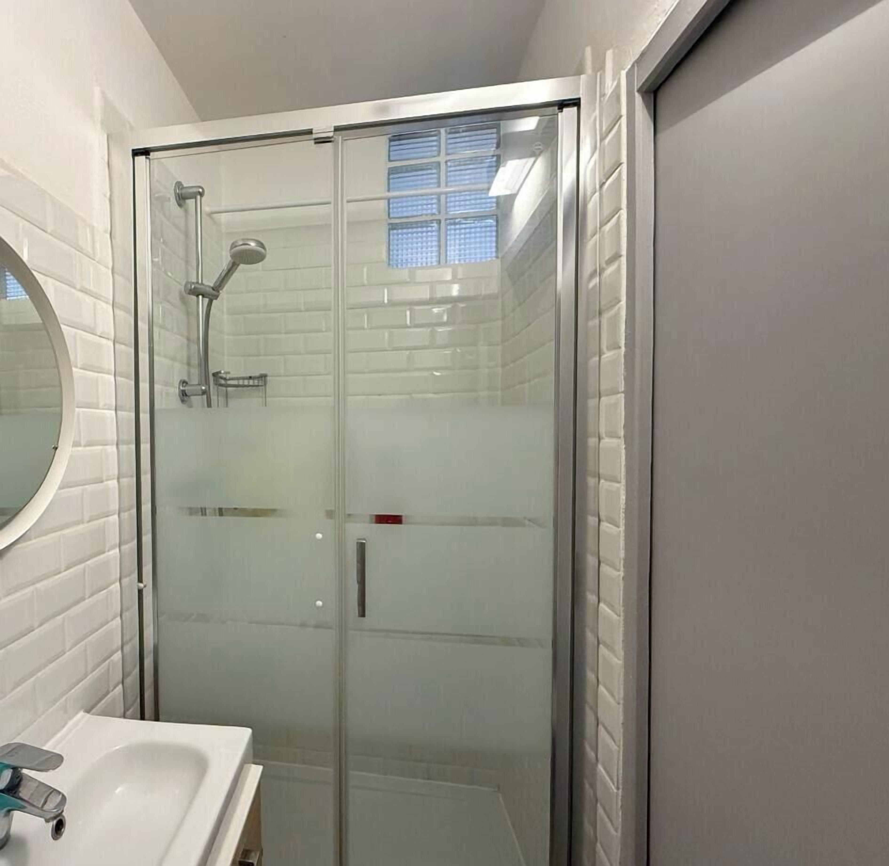 A glass-enclosed shower with a handheld showerhead, a glass door, and a white tiled wall.