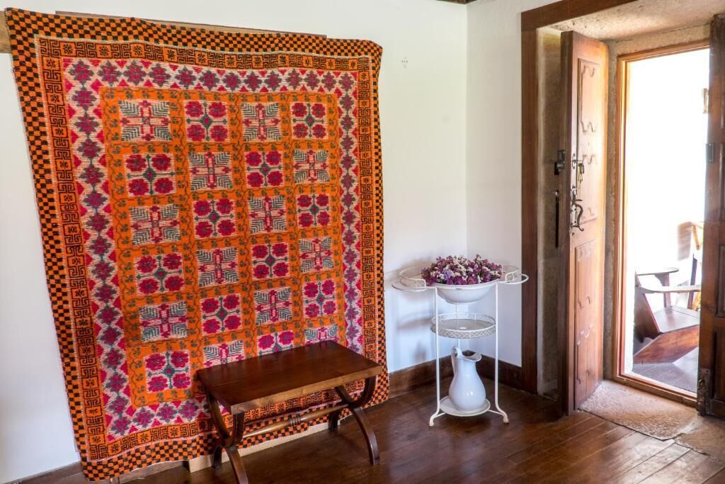 A room with a large quilt hanging on the wall, a small wooden table, and a white pedestal with a vase of flowers.