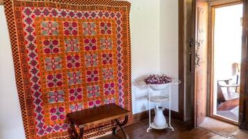 A room with a large quilt hanging on the wall, a small wooden table, and a white pedestal with a vase of flowers.