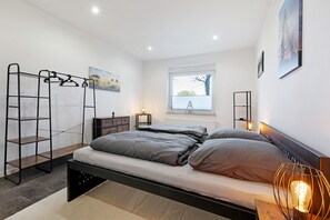 A modern bedroom with a bunk bed, a ladder, a bedside table with a lamp, a window with blinds, and a painting on the wall. - Revierruhe Apartment – Modern Comfort in Marl with Garden Views (Marl)
