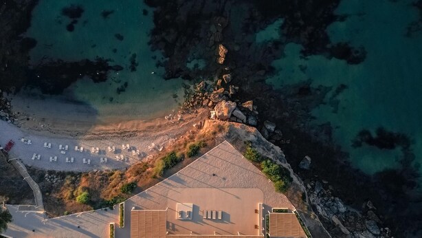 Eine Luftaufnahme einer Küstenimmobilie mit einer gepflasterten Terrasse, einem Strand mit Liegestühlen und felsigem Gelände.
