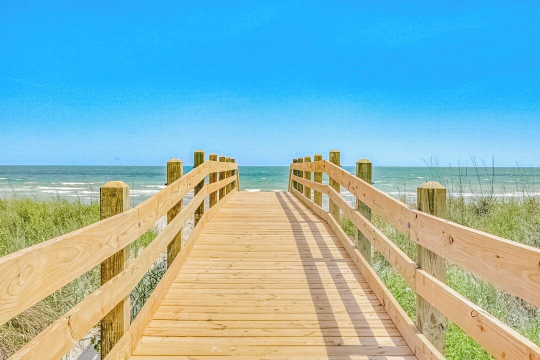Ein Holzsteg führt zum Strand, der Himmel ist klar.