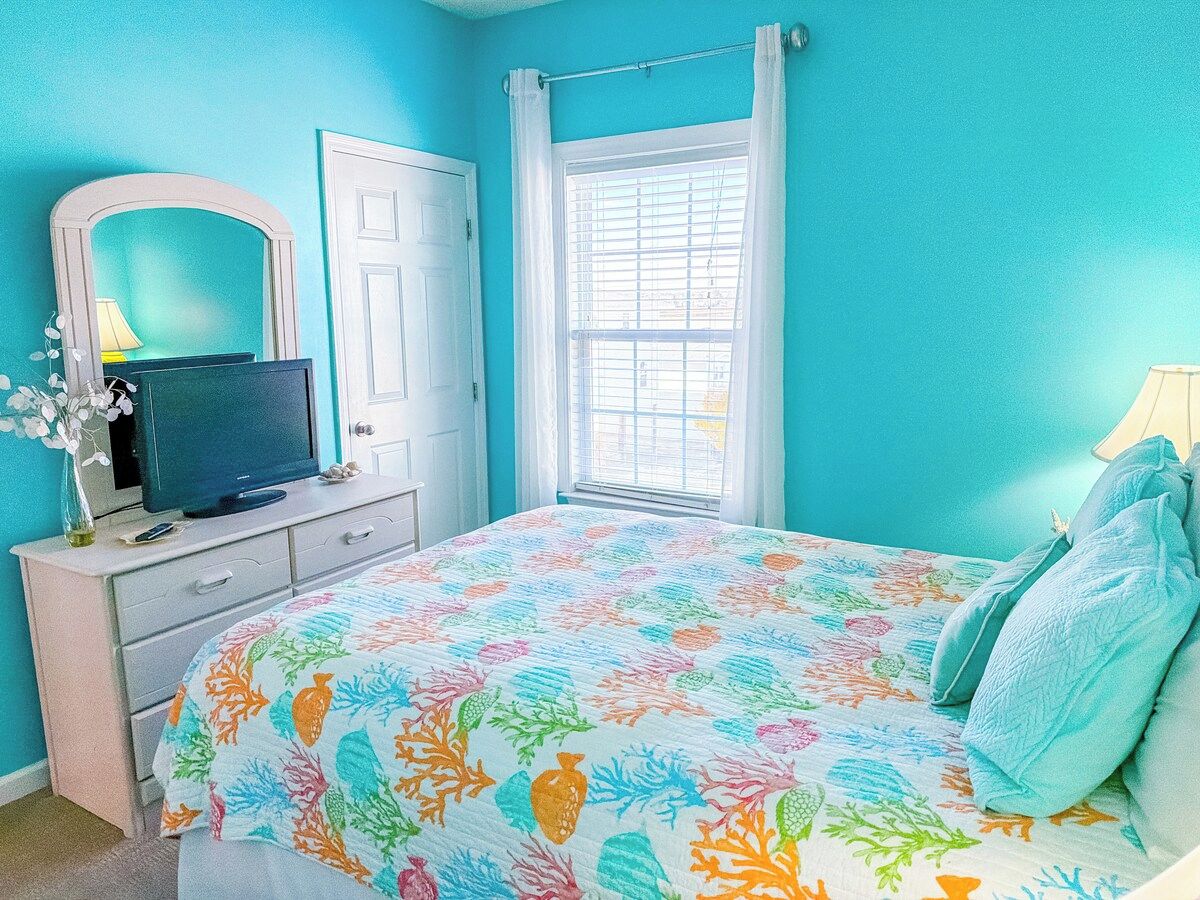 A bedroom with a bed, a dresser, a television, and a window with curtains.