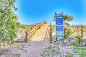 A wooden boardwalk leading to a beach area with a signboard. - Pelican Manor Unit D: 4 Bdrm/4.5 Ba, with wrap-around balconies with marsh views, modern décor, updated appliances, ample parking, and just a 1-minute walk to the beach. (North Myrtle Beach)