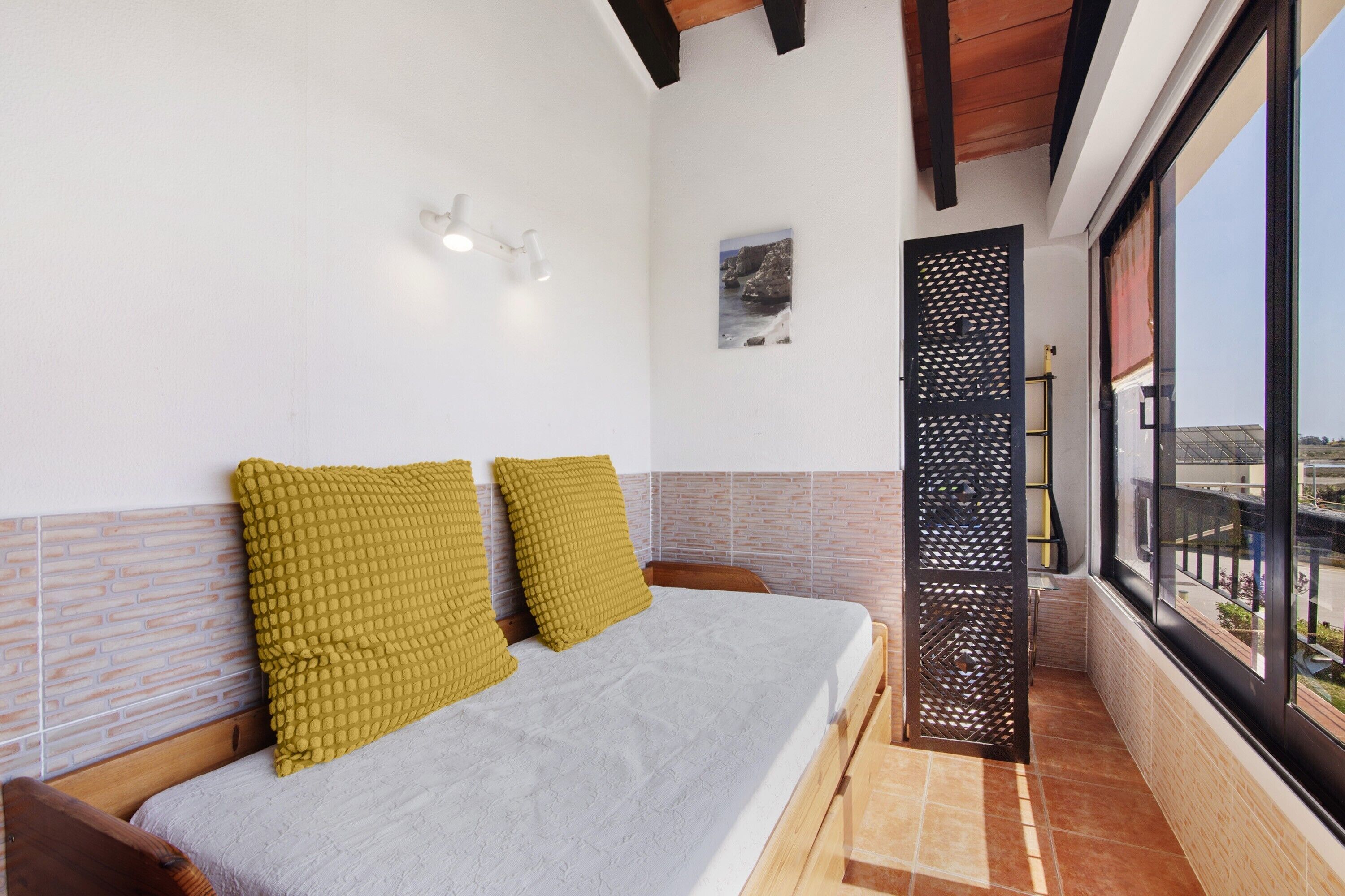 A bedroom with a bed, two pillows, a wall-mounted light, a picture, and a window with a view.