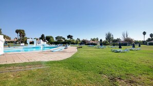 Piscina con sillas de descanso y sombrillas, rodeada de césped y árboles.