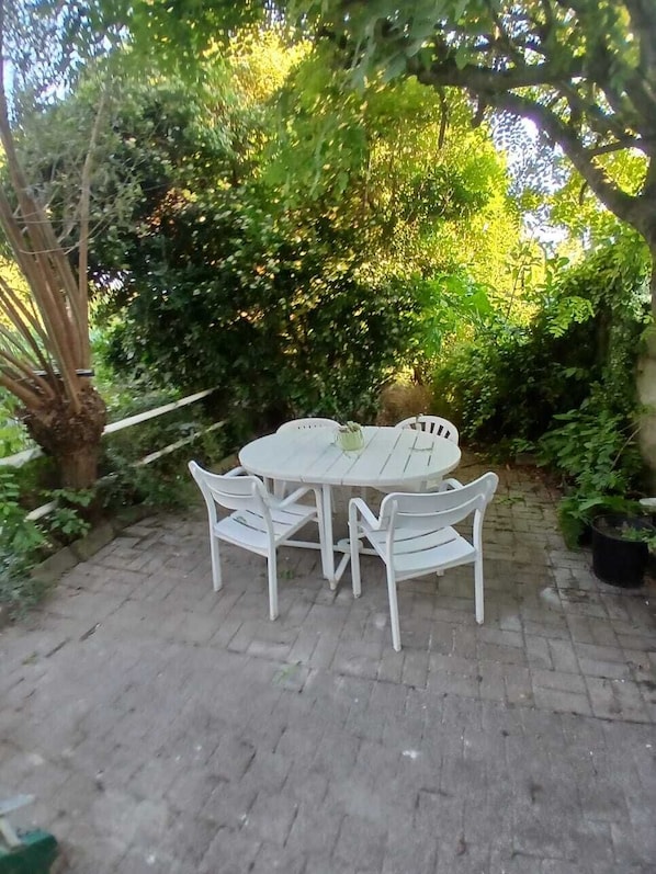 A round outdoor table with four chairs, surrounded by lush greenery and a brick pathway. - It's nice to meet you. (Asse)