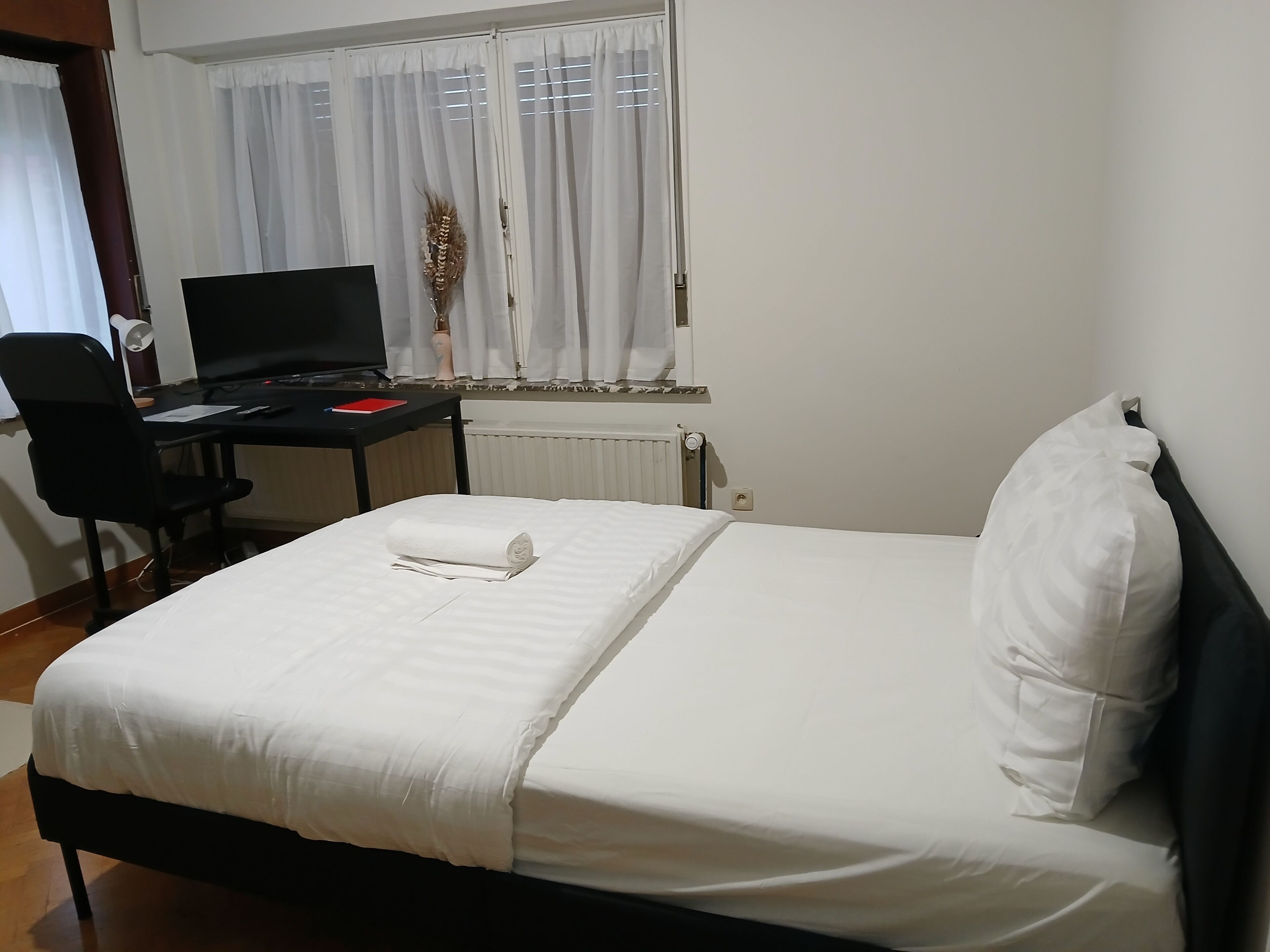 Quarto de hotel com uma cama, uma escrivaninha com computador, uma cadeira e uma janela com cortinas.