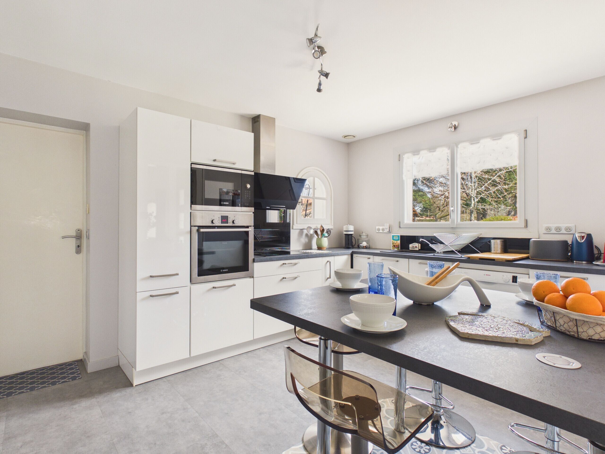 Una cocina moderna con armarios blancos, electrodomésticos de acero inoxidable, encimera negra y ventana con vistas a árboles.