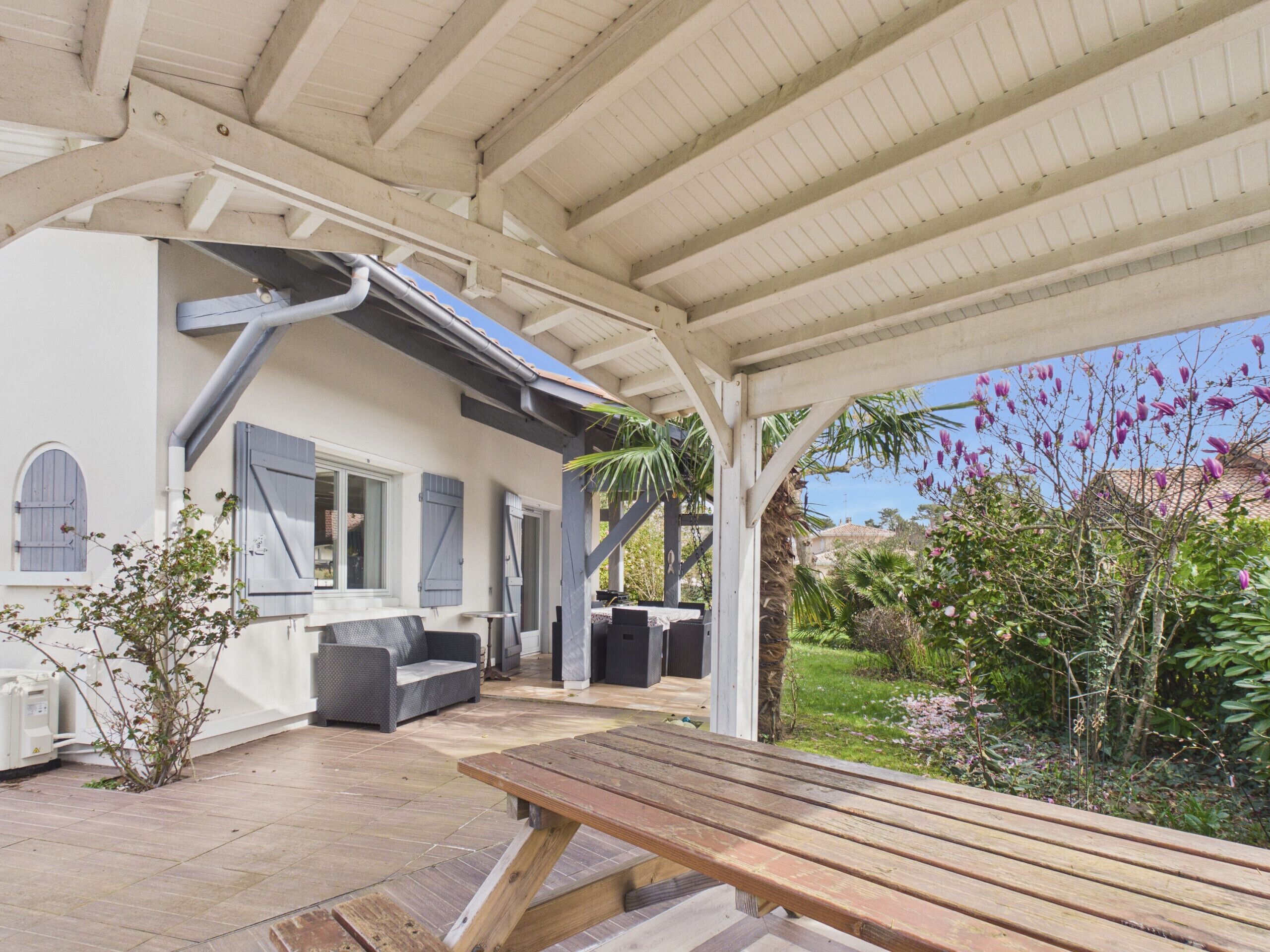 Un patio couvert avec un banc en bois, des sièges d’extérieur et une vue sur une maison à l’extérieur blanche et aux volets bleus.