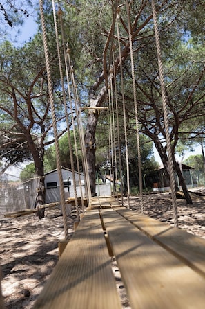Un puente colgante sobre una zona arenosa con árboles y un edificio pequeño al fondo.