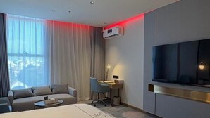 Una habitación de hotel moderna con cama, escritorio, silla, televisor y un ventanal grande con cortinas.