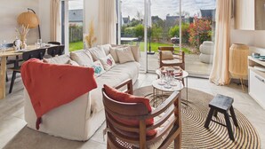 Ein modernes Wohnzimmer mit einer Couch, Sesseln, einem Esstisch und Blick durch große Glastüren auf einen Garten.