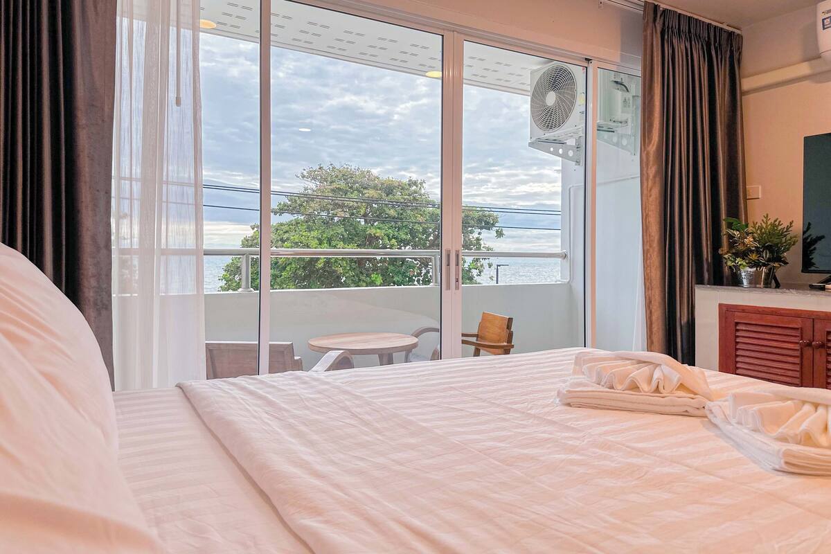 Una habitación de hotel con una cama grande, una mesita, una silla, un balcón con vista al mar y un ventilador de techo.