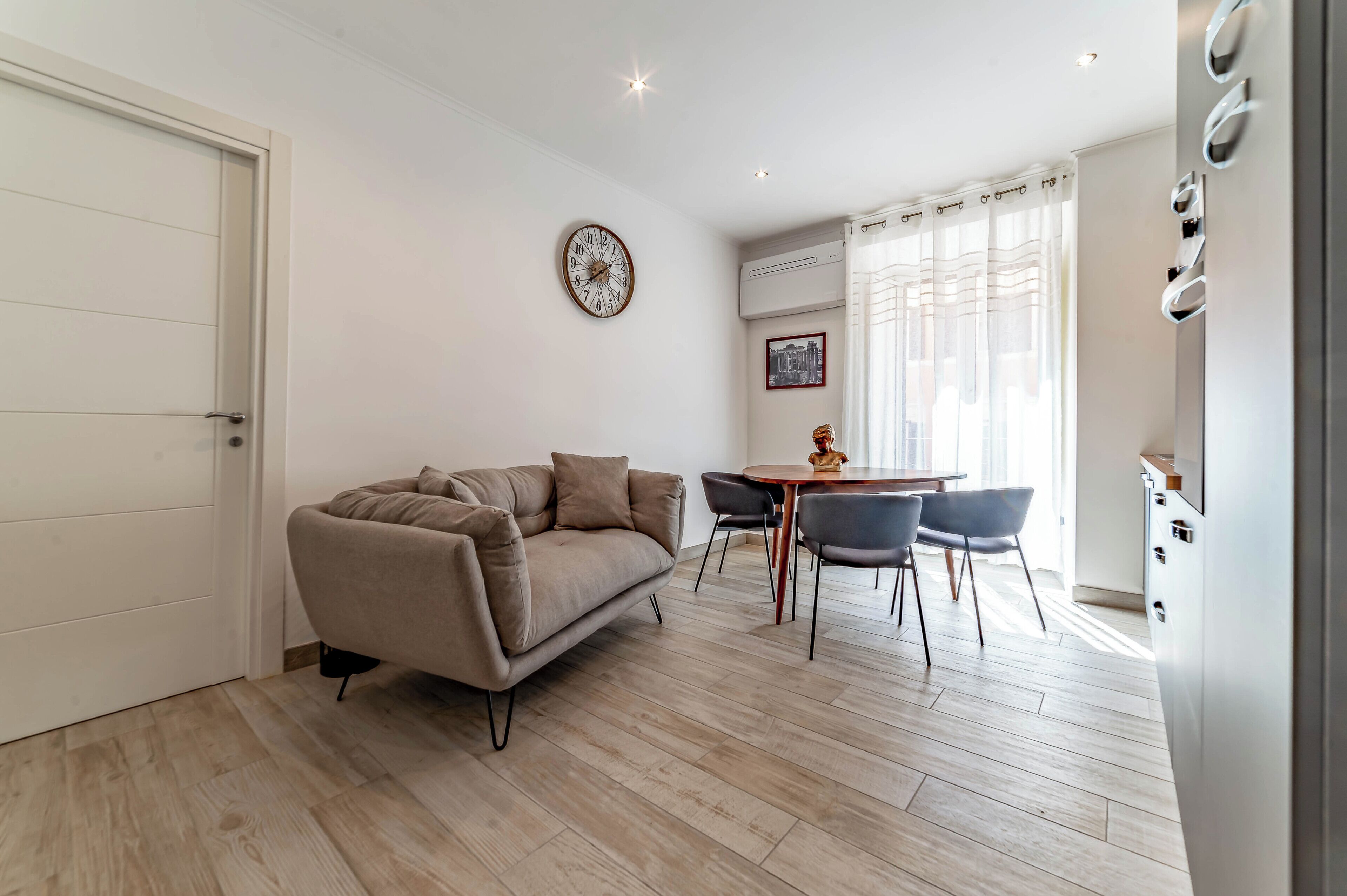 A modern living room with a sofa, dining table, chairs, a clock, and a kitchen area.