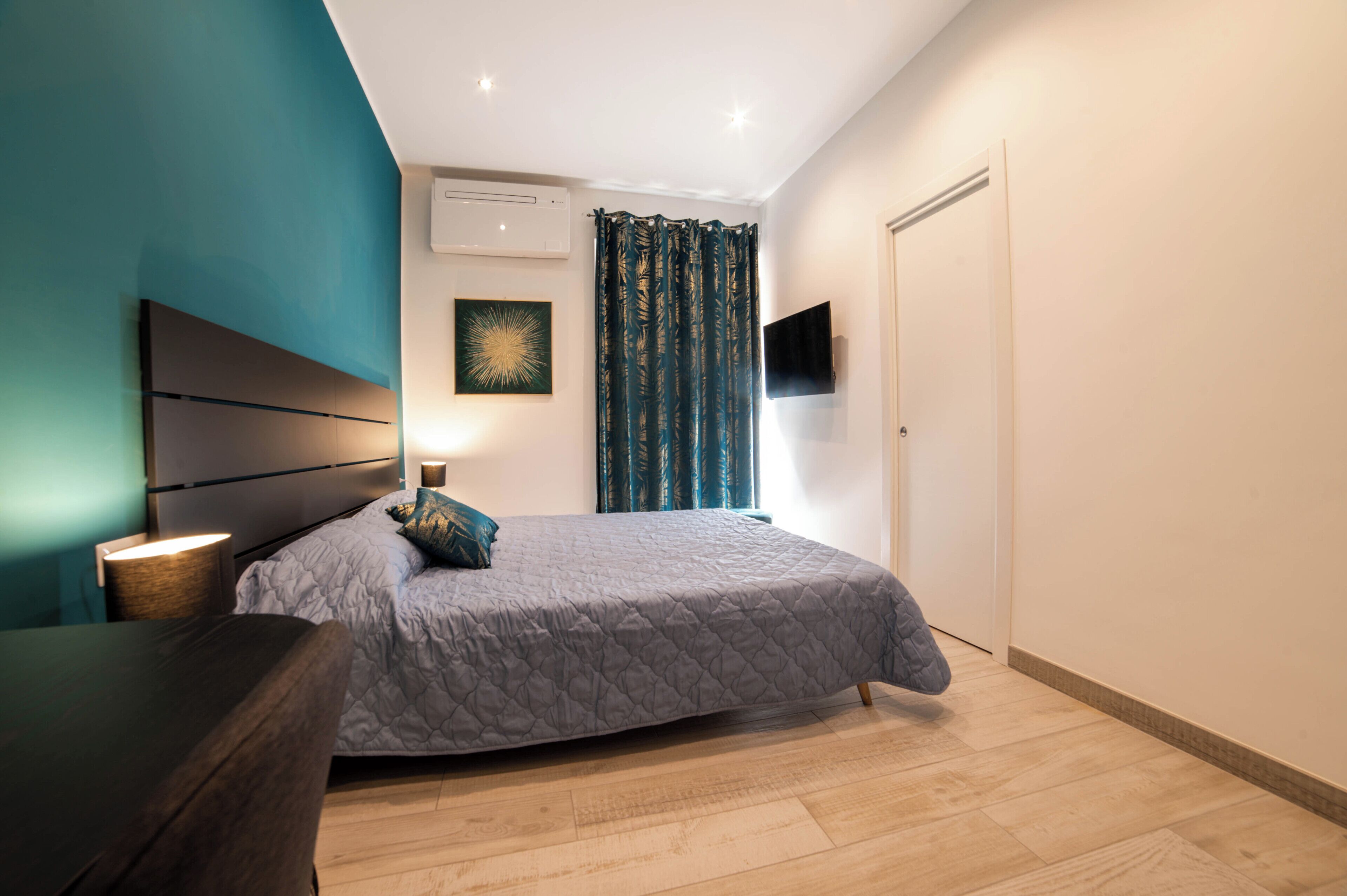 Une chambre à coucher comprenant un lit, une table de chevet, une lampe, un appareil de climatisation et un téléviseur fixé au mur.
