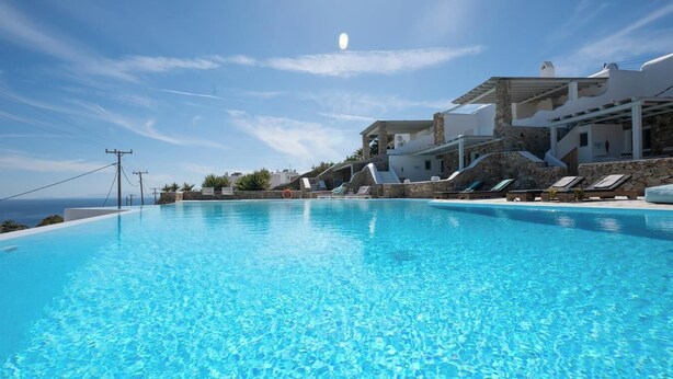 A clear blue infinity pool with lounge chairs and a stone building with a white facade. - Villa THEA Myconian Vision Villas (C9GQ+Q6 Ελιά, Greece)