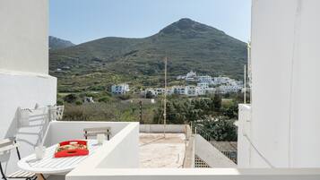 Un balcon avec une table, des chaises et une boîte rouge. En arrière-plan, on aperçoit des bâtiments blancs et une colline.