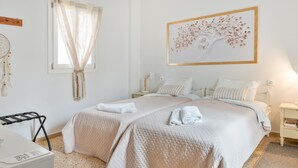 Een slaapkamer met twee bedden, een raam met dunne gordijnen, een aan de muur bevestigde droomvanger en een kunstwerk van een boom in een lijst.