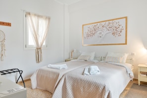 Un dormitorio con dos camas, una ventana con cortinas translúcidas, un atrapasueños colgado en la pared y una obra de arte con un árbol enmarcado.
