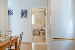 A kitchen with a white refrigerator, a dining table with chairs, and a hallway leading to a room with a bed and a window.