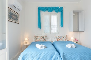 Ein Hotelzimmer mit zwei Betten, einem Fenster mit Vorhängen, einer Wand-Klimaanlage, einem Nachttisch mit Lampe und einem Spiegel.