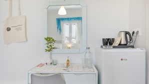 A small, clean room with a white desk, a mirror, a plant, a kettle, and a refrigerator.