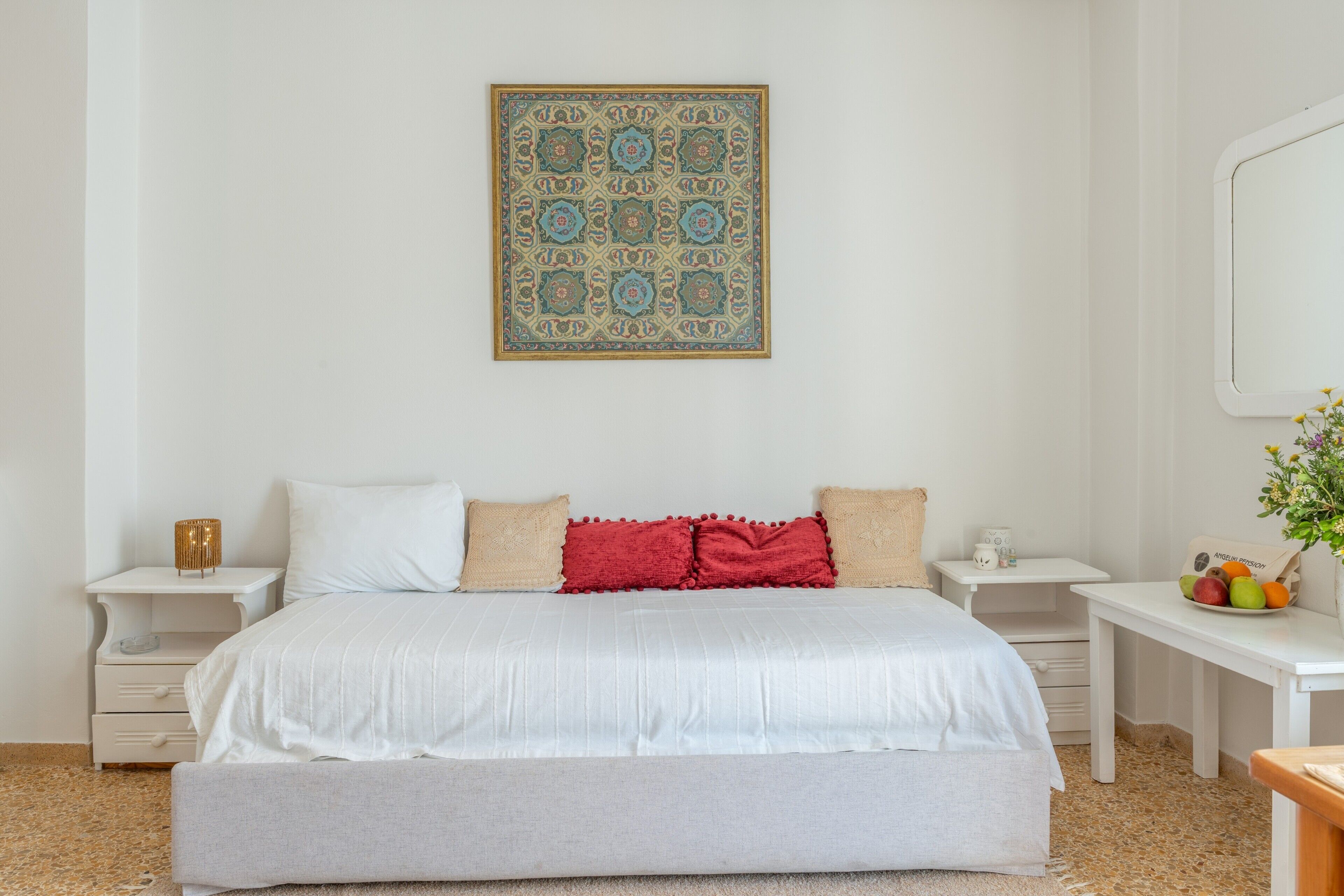 Un dormitorio con una cama grande, mesitas de noche blancas, una pequeña mesa con fruta y un adorno en la pared.