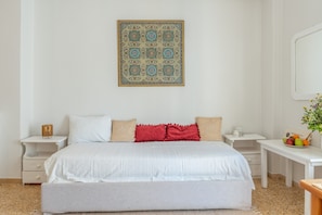 A bedroom with a large bed, white nightstands, a small table with fruit, and a wall hanging. - Studio Apartment with Balcony, Wi-Fi and Air Conditioning (Katápola)