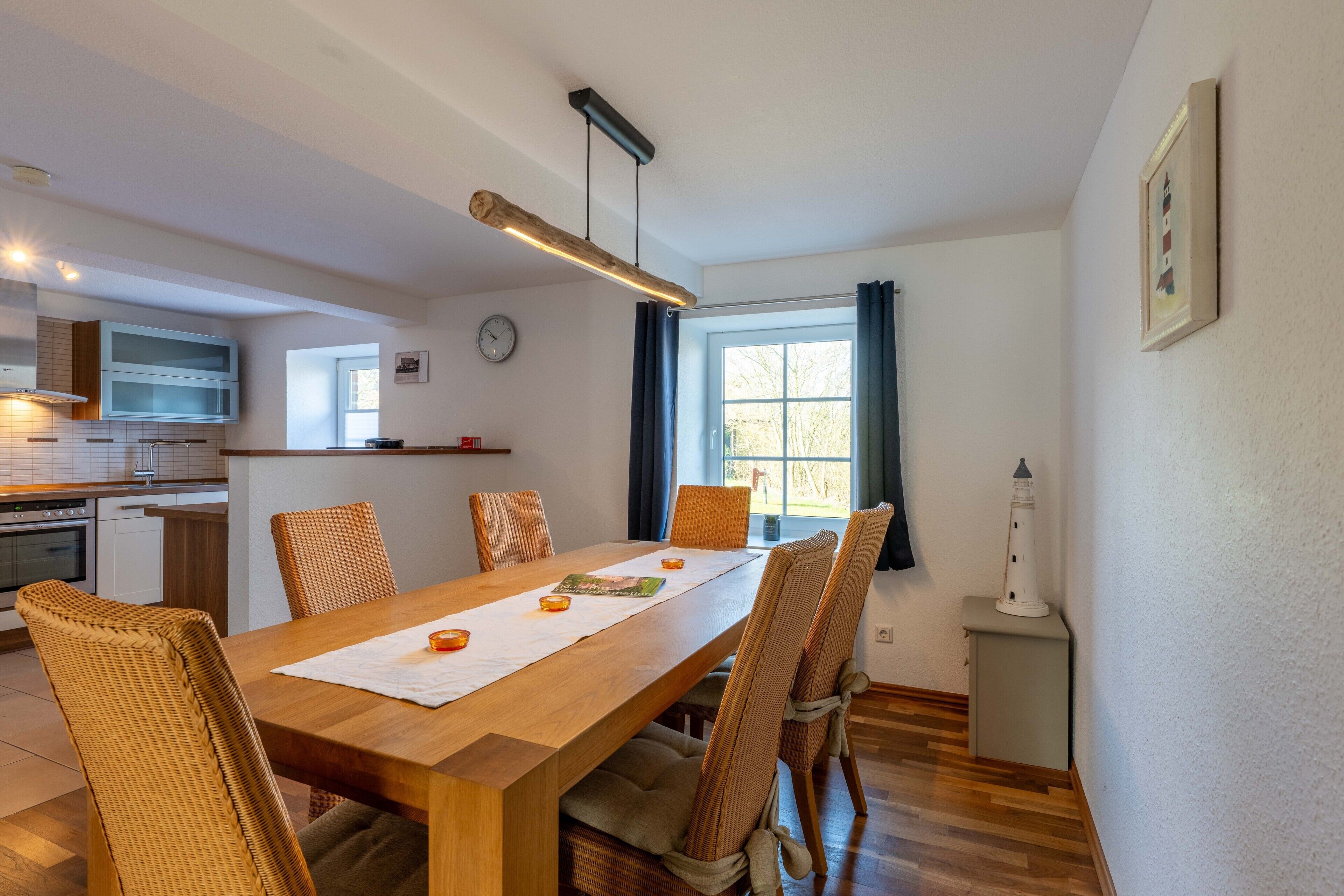 A dining area with a wooden table, wicker chairs, a pendant light, a small lighthouse decoration, and a view of the kitchen and living area.