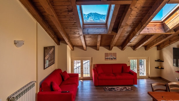 A spacious room with a high ceiling, wooden beams, a skylight, a red sofa, a dining table, and a view of mountains through the skylight. - Roncobellissimo! – Fresco e relax a 1.000 metri (Roncobello)