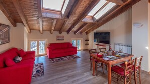 A spacious living area with a high wooden ceiling, a red sofa, a dining table set for four, and a large window providing natural light. - Roncobellissimo! – Fresco e relax a 1.000 metri (Roncobello)