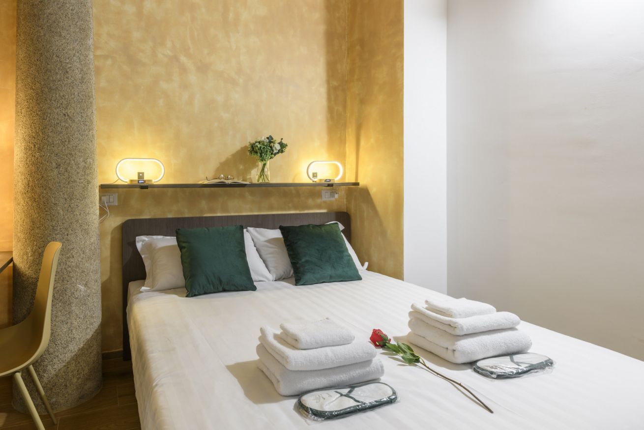 A neatly made bed with white linens, a vase with green foliage, and a rose on the bed.