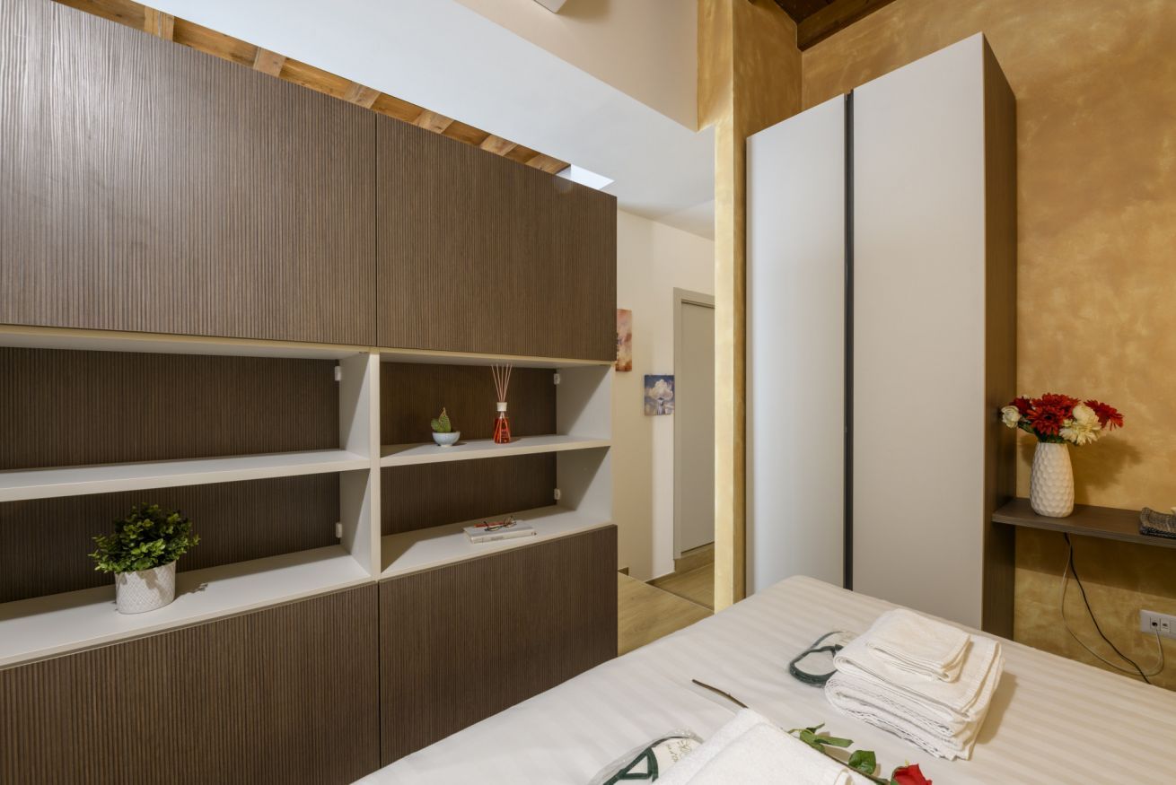 Una habitación de hotel con una cama, un escritorio con lámpara, un estante con adornos y un armario.