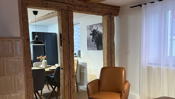 A living room with a brown leather chair, a wooden floor, a dining area with a table and chairs, and a kitchen in the background. - Premium Holiday Home - das1850, Historic, Modern, Deluxe (Balgheim)