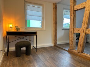 Une pièce avec un bureau en bois, un petit tabouret, une fenêtre avec des stores et une structure en poutres apparentes.