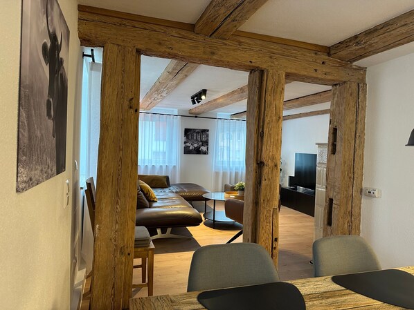 Un salon moderne avec des poutres apparentes, un canapé en cuir, une table à manger avec des chaises et un téléviseur.
