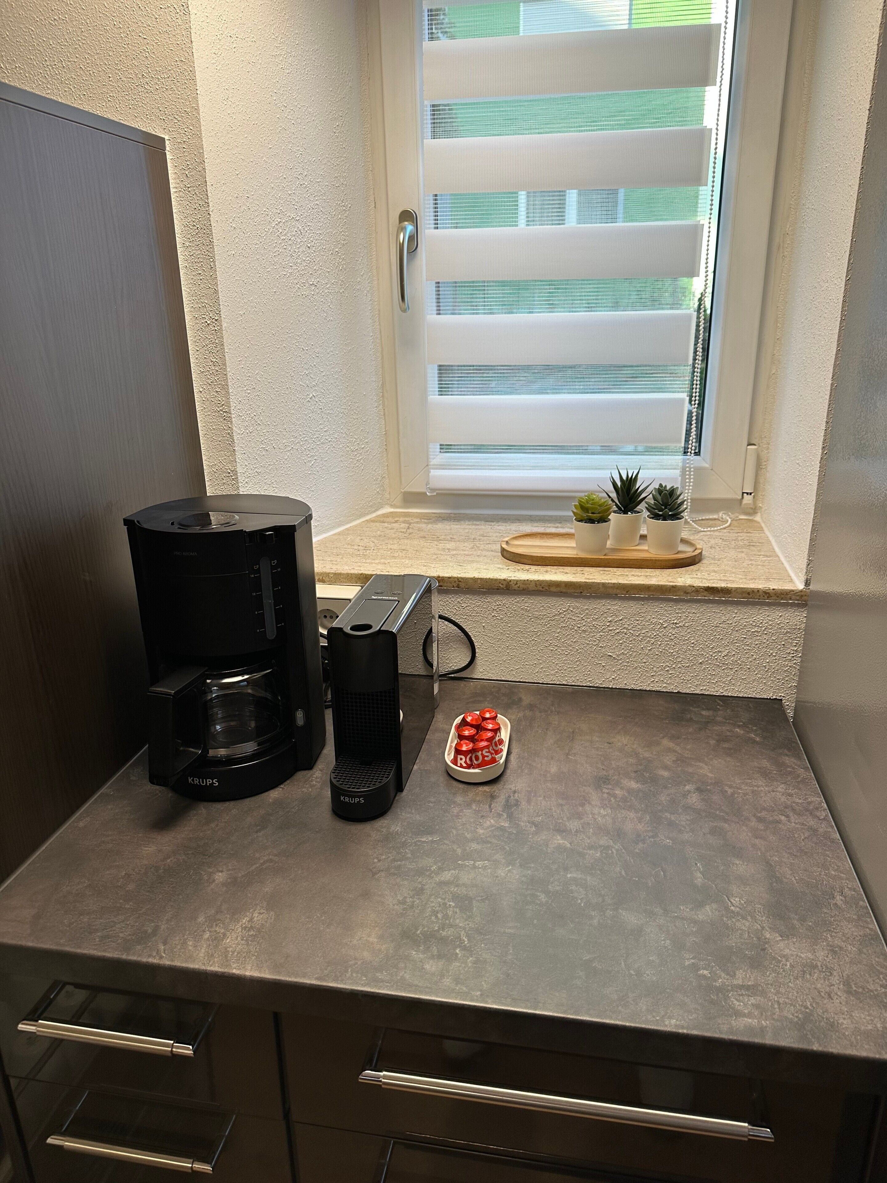 A kitchenette with a coffee maker, a small potted plant, and a tray of red objects.
