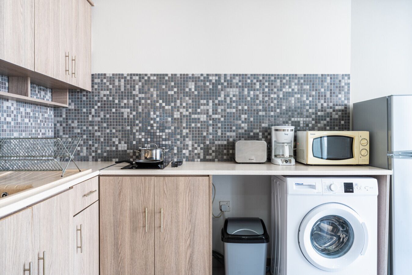 A compact kitchen with a microwave, toaster, and washing machine, a tiled backsplash, and wooden cabinets.