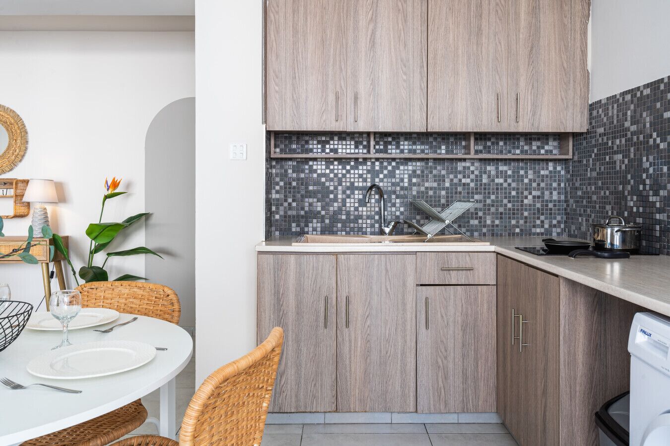 A modern kitchen with wooden cabinets, a tiled backsplash, and a dining area with wicker chairs and a table set for two.