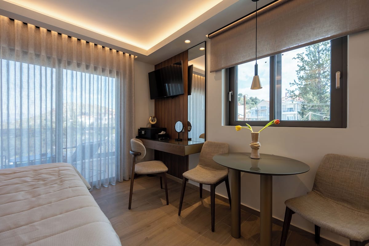 A modern hotel room with a bed, a desk with a chair, a round table, and a large window.