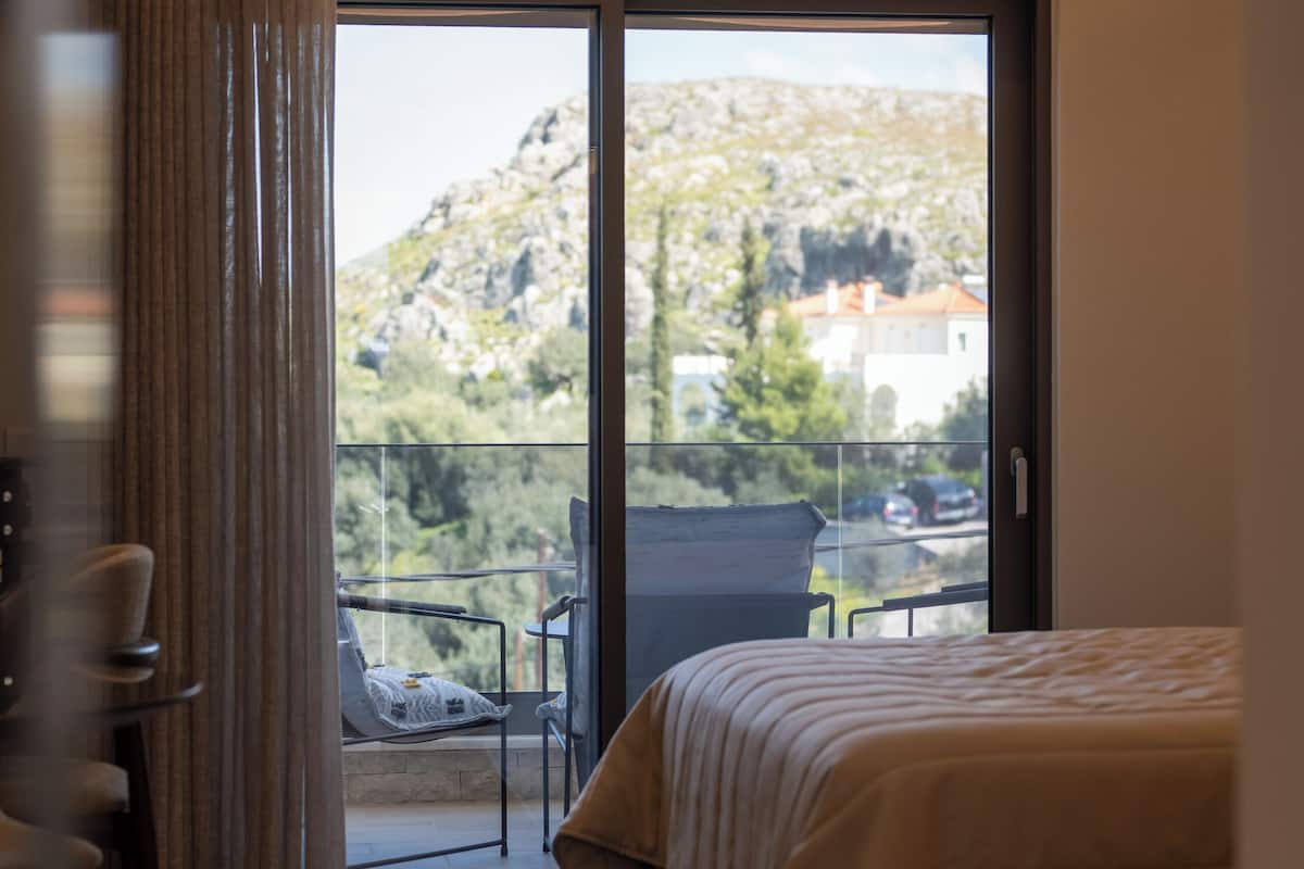 A hotel room with a bed, a small table, a chair, and a balcony with a view of the mountains.